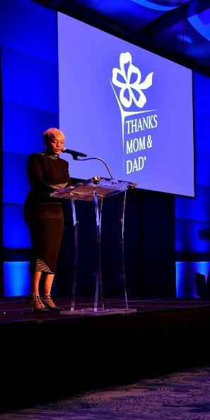 LaTasha speaking on a blue-lit stage beneath a Thanks Mom & Dad Fund screen